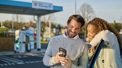 Un homme et une femme se tiennent souriants devant une borne de recharge Shell Recharge et regardent ensemble un smartphone. À l'arrière-plan, on aperçoit plusieurs bornes de recharge turquoise.
