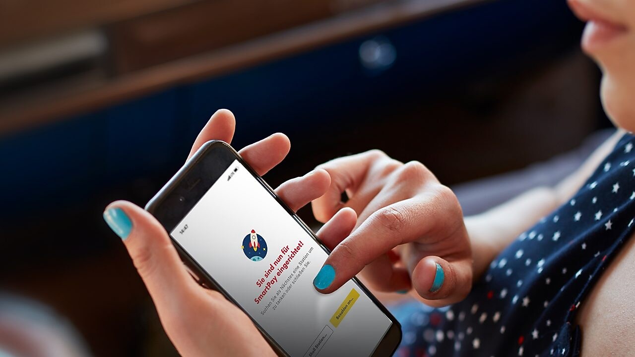 Une femme est assise dans sa voiture et tient un téléphone avec l’application Shell ouverte et Shell SmartPay configuré