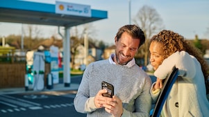 Ein Mann und eine Frau stehen lächelnd vor einer Shell Recharge Ladestation und schauen gemeinsam auf ein Smartphone. Im Hintergrund sind mehrere türkisfarbene Ladesäulen zu sehen.