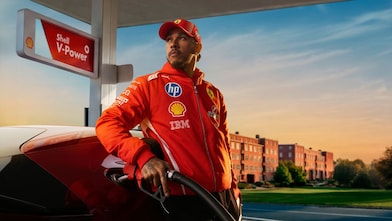 Formel-1-Fahrer Lewis Hamilton in rotem Rennanzug betankt einen roten Sportwagen an einer Zapfsäule mit Shell V-Power Beschilderung bei warmem Abendlicht.