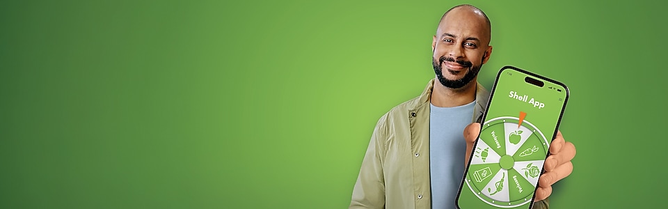 Ein dunkelhaariger junger Mann mit Bart vor einem grünem Hintergrund hält lächelnd ein Handy in die Kamera mit einem Glücksrad auf dem Display