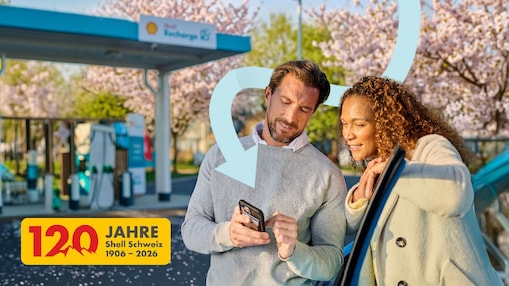 Ein Mann im hellgrauen Pullover und eine Frau im beigen Mantel lehnen an einem Auto an einer Shell Recharge Ladestation, schauen gemeinsam auf ein Smartphone, während hinter ihnen ein großer, geschwungener Pfeil und blühende Kirschbäume im Hintergrund zu sehen sind; im Vordergrund steht ein gelbes Jubiläumslogo mit der Aufschrift „120 Jahre Shell Schweiz 1906–2026“.