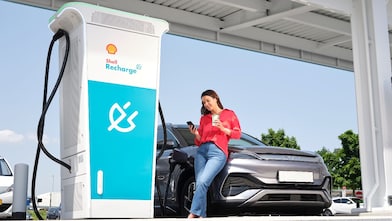 Eine Person benutzt ein Smartphone und lehnt sich an ein schwarzes Elektroauto, das an einer Shell Recharge Ladestation aufgeladen wird