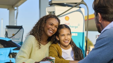 Eine Frau und ein Kind sitzen lächelnd an einem Tisch im Freien an einer Shell Recharge Ladestation. Ein Mann sitzt ihnen gegenüber. Im Hintergrund ist ein Elektrofahrzeug an einer Ladesäule zu sehen.