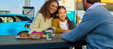 Eine Frau und ein Kind sitzen lächelnd an einem Tisch im Freien an einer Shell Recharge Ladestation. Ein Mann sitzt ihnen gegenüber. Im Hintergrund ist ein Elektrofahrzeug an einer Ladesäule zu sehen.