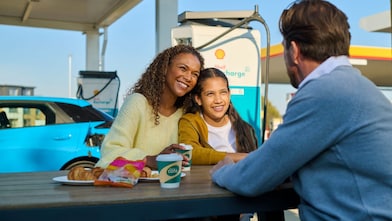 Eine Frau und ein Kind sitzen lächelnd an einem Tisch im Freien an einer Shell Recharge Ladestation. Ein Mann sitzt ihnen gegenüber. Im Hintergrund ist ein Elektrofahrzeug an einer Ladesäule zu sehen.