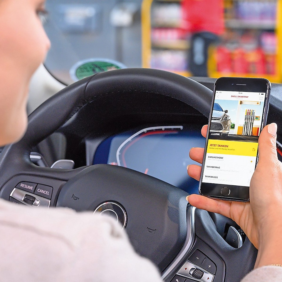 Blick über die Schulter einer Frau, die im Auto sitzt und ein Smartphone in der Hand hält. Der Fokus liegt auf dem Bildschirm, darauf ist die Shell App geöffnet. Im Hintergrund ist unscharf eine Shell Station zu erkennen.