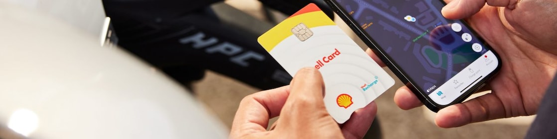 Ein Mann hält in der rechten Hand ein Smartphone, auf dem eine Karte abgebildet ist, in der linken Hand hält er eine Shell Card