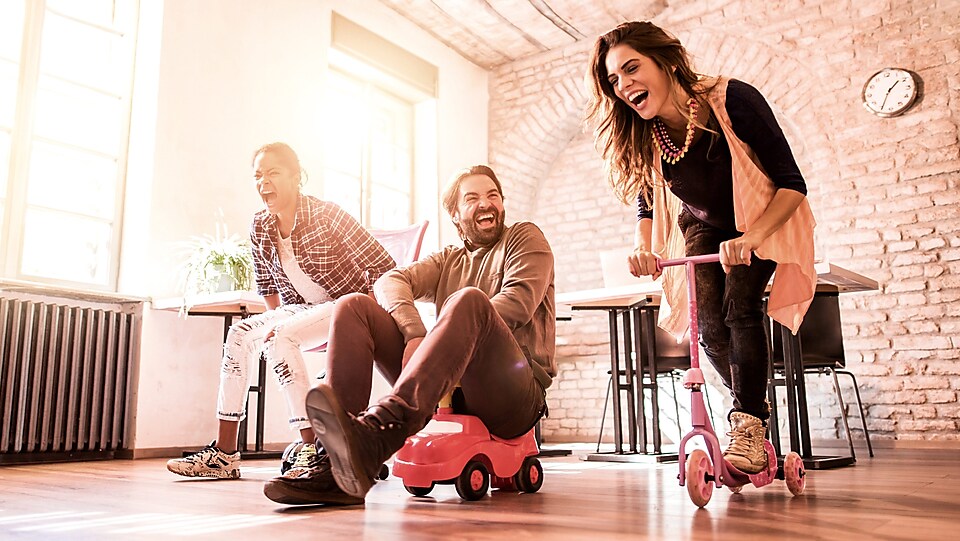 3 Personen fahren mit Spielzeugrollern und Spielzeugautos durch ein Zimmer.