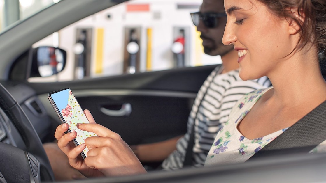 Eine Frau sitzt im Auto und hat auf dem Smartphone den Shell Card Stationsfinder offen. Neben ihr sitzt ein Mann.