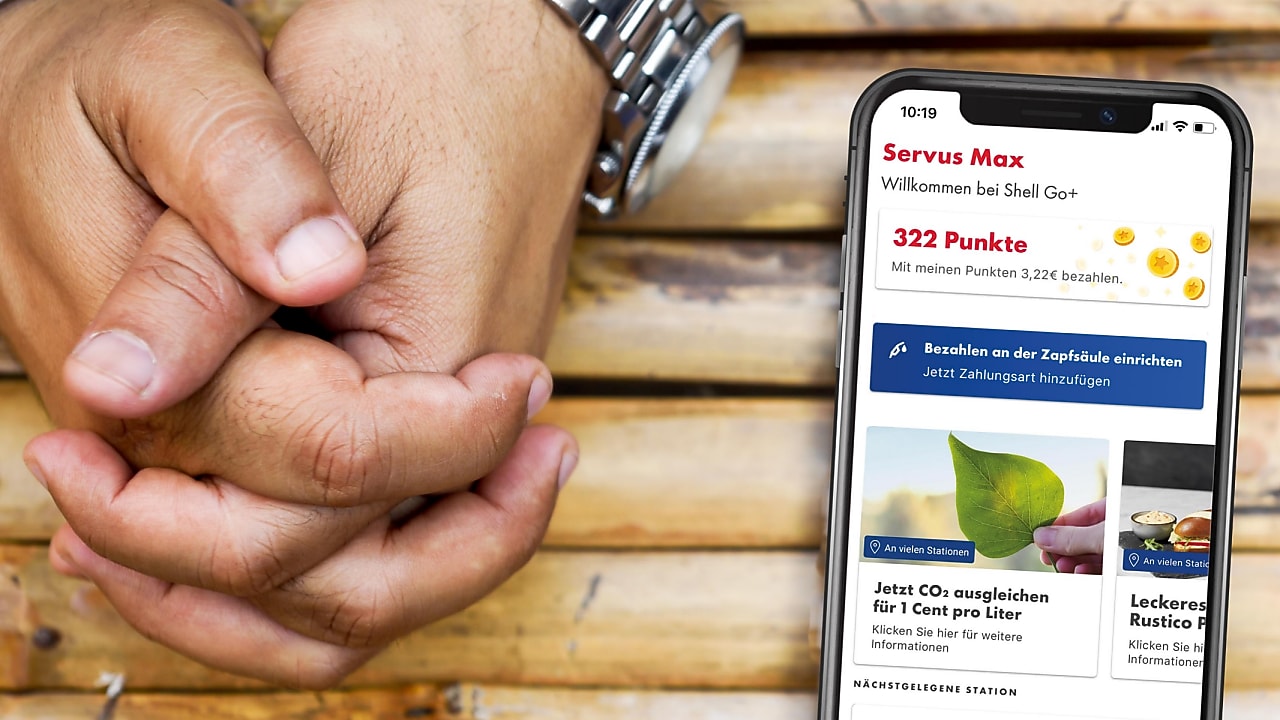 Smartphone mit Shell App, daneben gefaltete Hände auf Holztisch