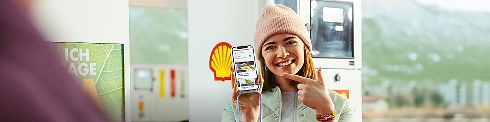 Hand mit Handy bricht durch eine gelbe Papierwand. Auf dem Display steht „Die neue Shell App %“. Links neben dem Handy ist ein roter Pfeil mit 4 Icons: Zapfpistole, Kaffeebecher, Waschanlage und drei Punkten (u.v.m.)