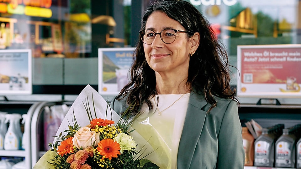 Eine dunkelhaarige Frau steht mit einem großen Blumenstrauß vor dem Tankstellen-Shop