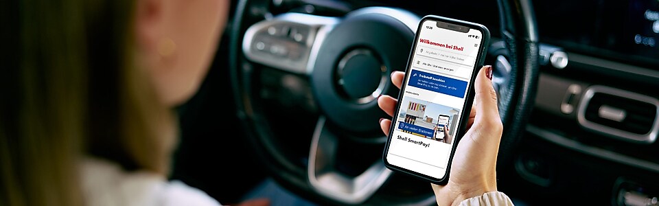 Shell App Startbildschirm auf Handy mit Tankstelle im Hintergrund