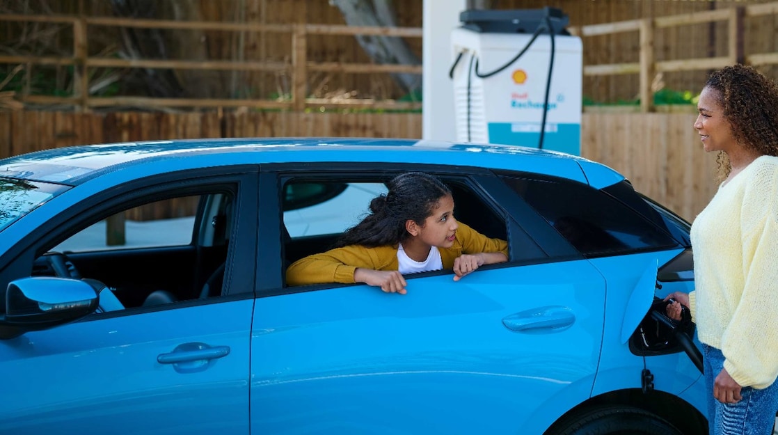Frau lädt ein blaues Elektroauto an einer Shell Recharge Ladesäule, während ein Kind aus dem geöffneten Autofenster schaut.