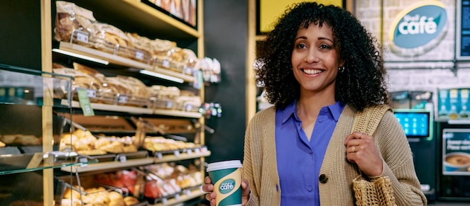 Frau steht lächelnd mit einem Kaffee von Shell Café in einer Shell Station mit Shell Café Backshop