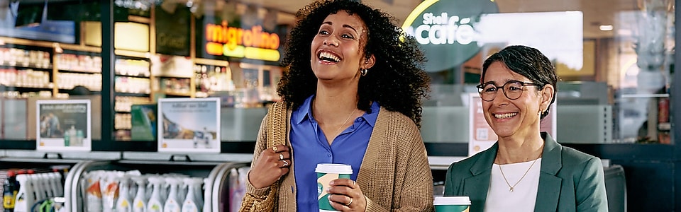 In einem Shell Cafè trinkt ein Mann Shell Café au einem Becher. Er lacht und hat einen Milchschaum um den Mund.