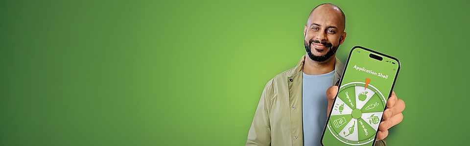 Ein dunkelhaariger junger Mann mit Bart vor einem grünem Hintergrund hält lächelnd ein Handy in die Kamera mit einem Glücksrad auf dem Display