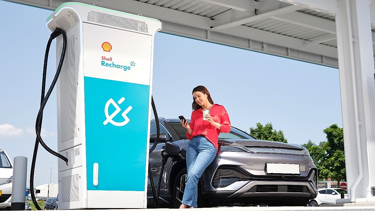 Une personne utilise un smartphone et s’appuie sur une voiture électrique noire qui est en train d’être rechargée à une borne Shell Recharge