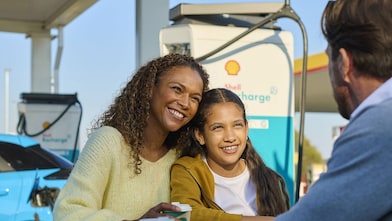 Une femme et un enfant sont assis à une table en plein air près d'une borne Shell Recharge, un sourire aux lèvres. Un homme est assis en face d'eux. À l'arrière-plan, on aperçoit un véhicule électrique branché sur une borne de recharge.