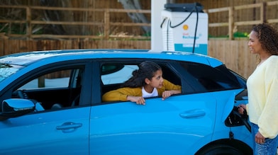 Une femme recharge une voiture électrique bleue sur une borne Shell Recharge, tandis qu’un enfant regarde par la fenêtre ouverte de la voiture.