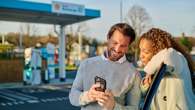 Un homme et une femme se tiennent souriants devant une borne de recharge Shell Recharge et regardent ensemble un smartphone. À l'arrière-plan, on aperçoit plusieurs bornes de recharge turquoise.
