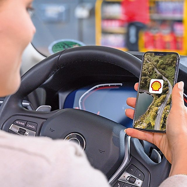 La photo montre la vue par-dessus l'épaule d'une femme assise dans une voiture et tenant un smartphone dans sa main. L'accent est mis sur l'écran, sur lequel l'application Shell est ouverte. À l'arrière-plan, on peut voir une station Shell hors champ.