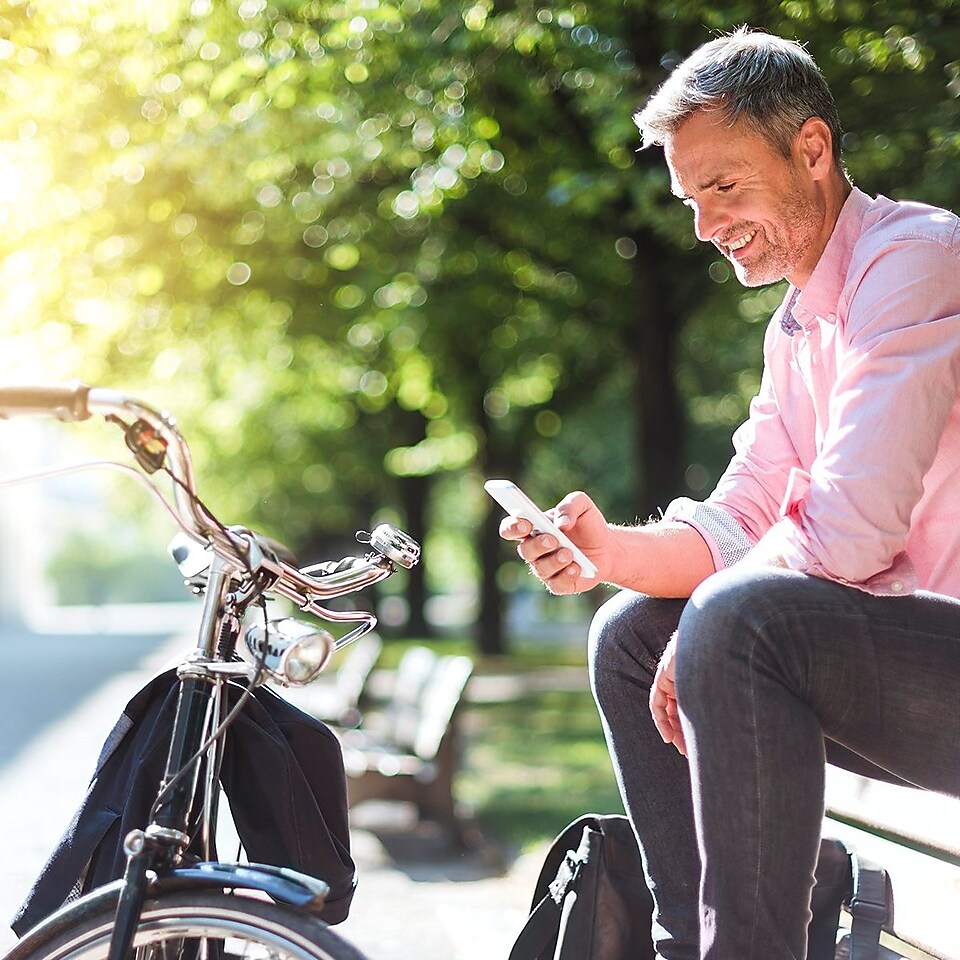 Un gestionnaire de flotte est assis, détendu, sur un banc de parc, son smartphone à la main. Devant lui se trouve sa bicyclette.