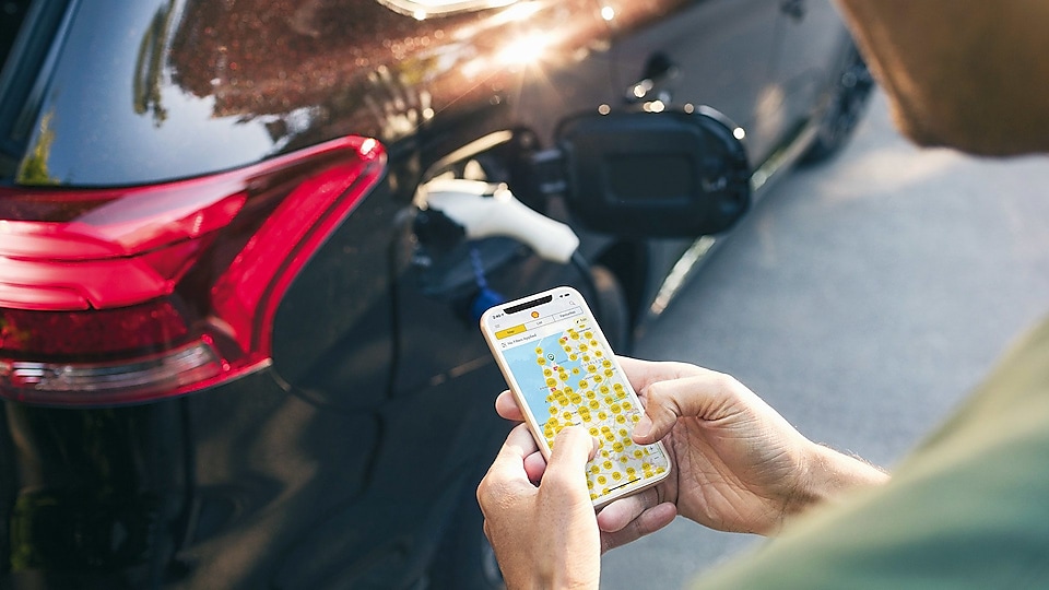 Au premier plan, un conducteur utilise le smartphone avec l'application NewMotion pour afficher les points de recharge disponibles. En arrière-plan, le véhicule électronique se recharge à une station de mobilité électronique.