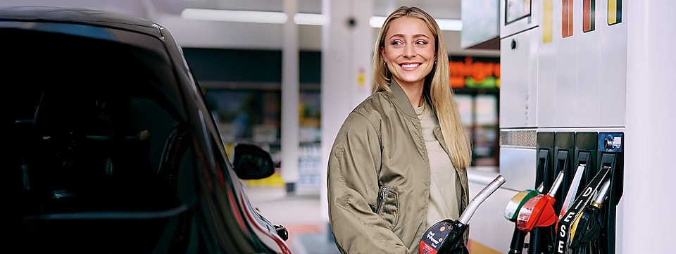 Une jeune femme blonde sourit devant une pompe à essence Shell, un pistolet à la main.