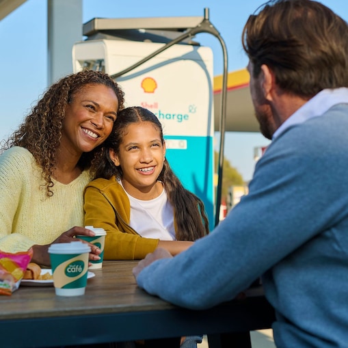Femme Une femme et un enfant sont assis à une table en plein air près d'une borne Shell Recharge, un sourire aux lèvres. Un homme est assis en face d'eux. À l'arrière-plan, on aperçoit un véhicule électrique branché sur une borne de recharge.utilisant une carte de recharge à une station Shell Recharge