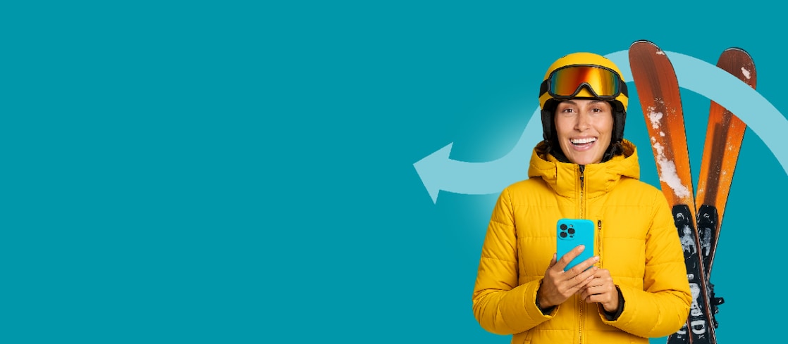 Femme souriante en veste de ski jaune avec casque et masque de ski tenant un smartphone, devant un fond turquoise.