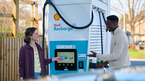 Une femme et un homme debout devant une borne de recharge Shell Recharge. La femme utilise l'écran tactile de la borne. En arrière-plan, on aperçoit un véhicule.