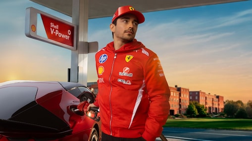 Le pilote de Formule 1 Charles Leclerc, en combinaison rouge, fait le plein d’une voiture de sport rouge à une pompe Shell V‑Power dans une lumière chaude de fin de journée.