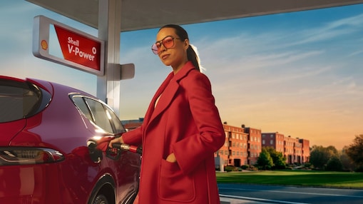 Femme en manteau rouge et lunettes teintées faisant le plein d’une voiture de sport rouge à une pompe avec signalisation Shell V‑Power dans une lumière chaude de fin de journée.