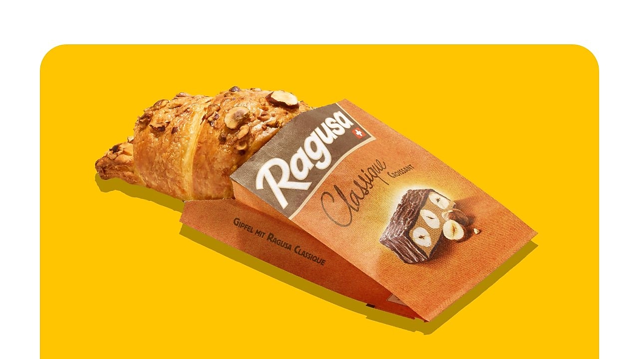 Croissant fourré au praliné dans un sachet en papier, inscription « Ragusa Classique Croissant » et « Gipfel mit Ragusa Classique » sur fond jaune.